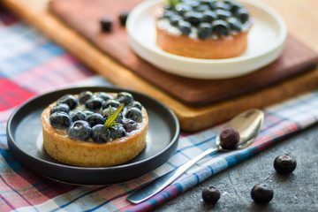 Delicious blueberry tartalets with vanilla cream on black and white plates on a cage napkin and a wooden board with a spoon on a grey background. Top view. Top view.