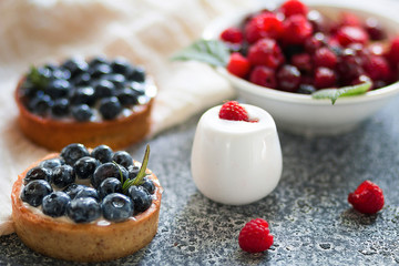 Tartalets with blueberry berries, plate with fresh raspberry and dairy with sour cream on the grey surface. 