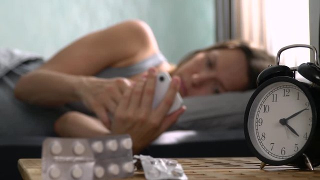 A lonely sick girl looks at her cell phone, counting the minutes until the pain subsides. A woman lies on the bed and hurts. There are Antidepressants on the bedside tables. Virus Allergy Infection
