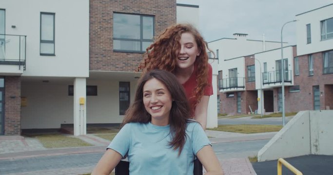 Pretty ginger girl riding her best friend in wheelchair outdoor. Attractive smiling invalid girl enjoying spare time on sunny day in modern city.
