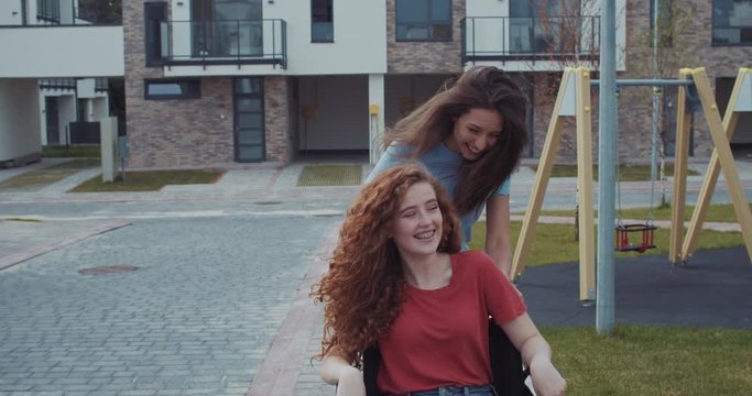 Attractive friendly girls having fun leisure time together outdoor. Happy friend riding disabled ginger smiling girl in wheelchair in the playground in modern city. Friendship concept.