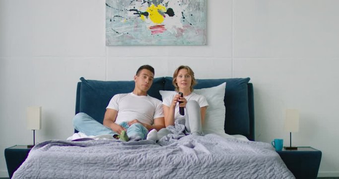 Young mixed couple is watching TV, relaxing in bedroom, woman is cheerful, watching her favorite TV show, film, holding remote controller, man is bored and in bad mood. Latin, white, 4K, RED camera.