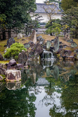 Fototapeta na wymiar Old mossy stones reflect in a dark green pond with a white Japanese tea house behind some trees and a fence