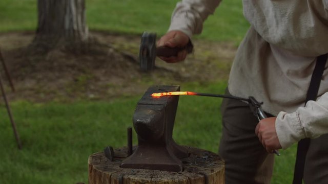 A revolutionary war reinacter blacksmith hammers out a hot piece of molten metal out on an anvil