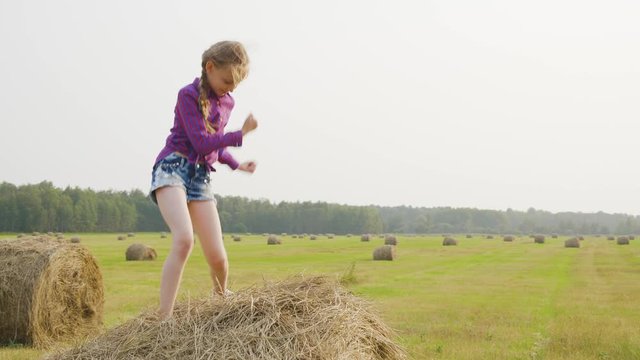 Happy girl dancing on haystack at harvesting field. Cheerfullteenager girl dancing on hay stack on countryside field. Young girl having fun on autumn field