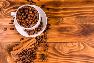 White cup filled with coffee beans, star anise and cinnamon sticks on wooden table. Top view