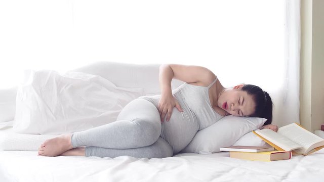 Beautiful asian pregnant having labor pain while lying to read a book on bed in bedroom