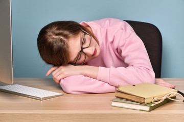 A girl in a pink sweater sits at a table with a computer and books with her head resting on folded...