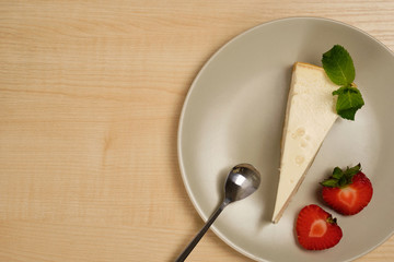 A plate with cheesecake, a spoon and strawberries stands on a light wooden table.