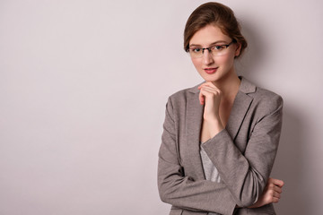 A girl in a gray jacket and glasses stands on a white background, looks at the camera, propping her...
