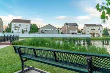 Green metal park bench with view of a grassy pond and homes under cloudy sky