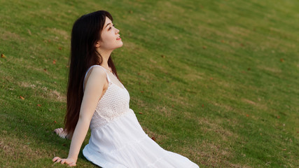 Fototapeta na wymiar Portrait of beautiful Chinese young woman in white wedding dress sitting on grass field with hand on ground, beauty in summer.