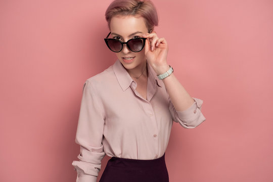 A girl with short pink hair in a blouse and skirt stands on a pink background, looking over sunglasses.