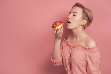 A girl with short pink hair in a blouse reaches for with a closed eyes and open mouth to a donut in her hand, on a pink background