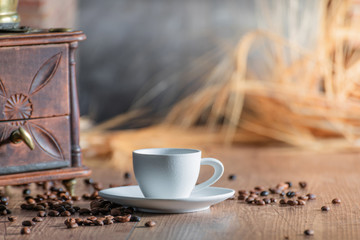 Spike of wheat and a small red mill behind a porcelain cup with coffee beans scattered on a wooden table. Relaxation concept and coffee aroma.