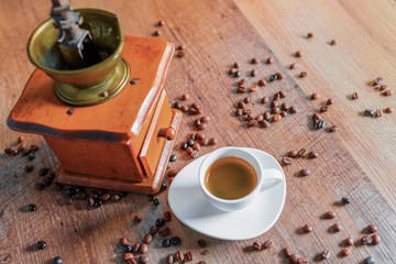 Concept of coffee aroma and relaxation in the morning. A small red mill next to a white cup of espresso coffee with many coffee beans scattered on a wooden table.