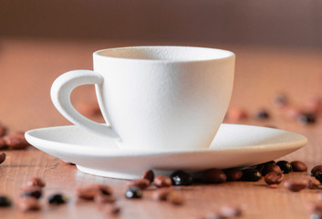 A white cup with a selective focus surrounded by large roasted coffee beans on a wooden table.