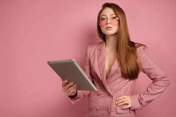 Fototapeta premium Red-haired girl in a jacket stands on a pink background with an electronic tablet and looks into the camera over glasses.