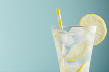 Refreshing summer tonic water with lemon, ice cubes, yellow straw in light soft pastel blue color background, closeup, half, top, edge.