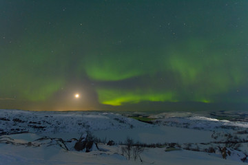 Northern lights, aurora in the sky at night. Hills and rocks covered with snow. Moon in the sky.