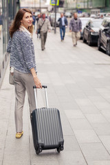 Fototapeta na wymiar vertical portrait of beautiful smiling elegant young woman in business clothes walks along the street along a glass building and rolls a small suitcase on wheels