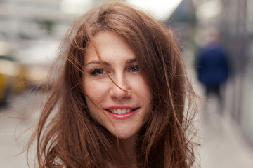 Obraz premium Close portrait of elegant beautiful young smiling brunette woman with messy long hair scattered by the wind standing on the street