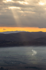 Sun rays coming down from some clouds over a valley filled by fog, illuminating part of it