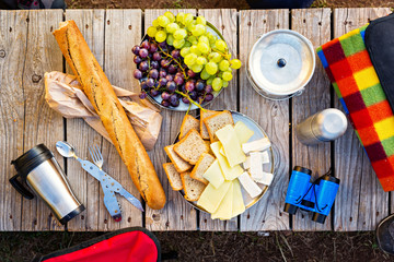 Top view of small snack in the mountains. Cheese, grapes and baguette bread next to camping equipment, on top of wooden table.
