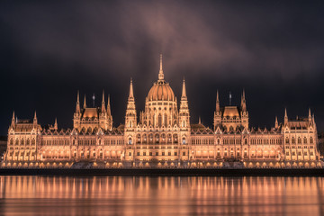 Illuminated Budapest parliament building at night with dark cloudy sky and reflection in Danube river