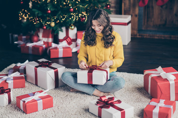 Photo of charming nice cute girl wearing jeans denim yellow sweater unpacking her new year gift from santa claus while sitting on carpet on floor with legs crossed - obrazy, fototapety, plakaty