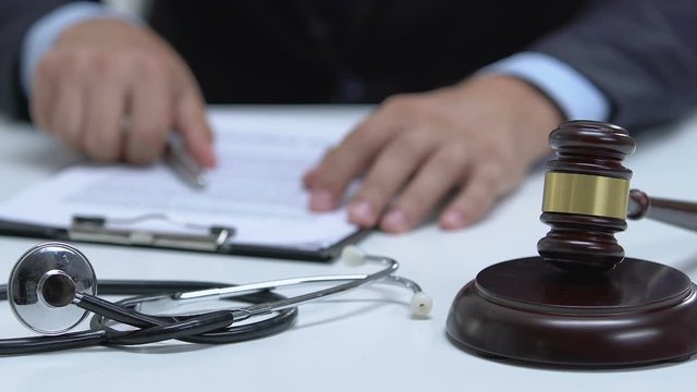 Male judge signing documents and taping gavel, stethoscope on table, negligence