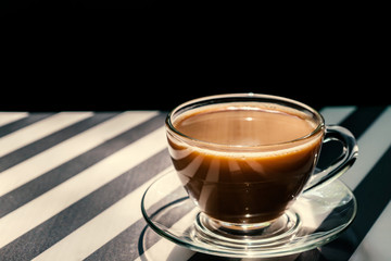 cup of coffee with milk on black and white striped backeound. Rays of light in cup. Copy space