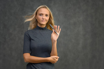 Obraz premium Studio portrait of a pretty blonde student girl, young woman in a gray sweater with a pen in her hands on a gray background. Talking, showing emotions.
