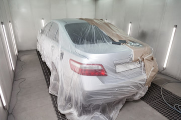 A large silver sedan car is completely covered in paper and adhesive tape to protect against splash during painting after an accident in a workshop for body repair of vehicles. Auto service industry.