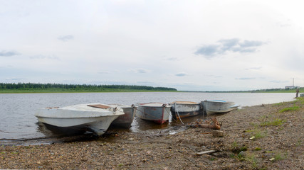 Fototapeta na wymiar Five old metal boats are tethered near the shore against the background of a girl fisherman and the village of Northern Yakutia in the taiga Suntar.