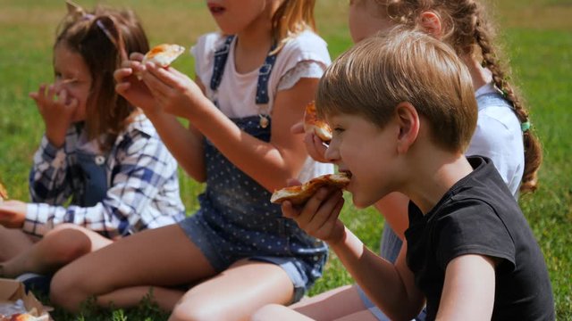 Group of children friends picnic eat pizza sit on park grass chewing and communicating