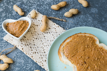 slice of bread with peanut butter, heart shape bowl and peanuts, on grey table