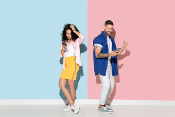Young emotional caucasian couple in bright casual clothes posing on pink and blue background. Concept of human emotions, facial expession, relations, ad. Woman and man using smartphone, happy, busy.