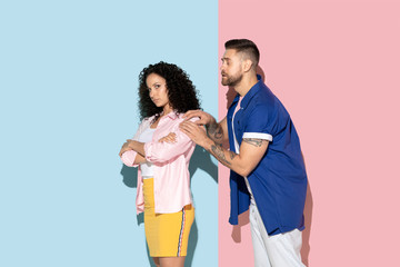 Young emotional caucasian couple in bright casual clothes posing on pink and blue background. Concept of human emotions, facial expession, relations, ad. Woman is offended, man apologizes.
