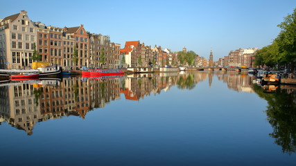 Crooked and colorful heritage buildings and houseboats, overlooking Amstel river with perfect reflections, with Munttoren (historic tower with a carillon) in the background, Amsterdam, Netherlands