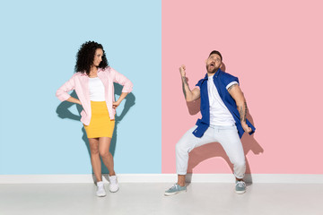 Fototapeta na wymiar Young emotional caucasian couple in bright casual clothes posing on pink and blue background. Concept of human emotions, facial expession, relations, ad. Man's dancing and celebrating, woman is bored.