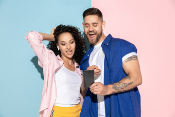 Young emotional caucasian couple in bright clothes posing on pink and blue background. Concept of human emotions, facial expession, relations, ad. Man and woman make selfie in wireless earphones.
