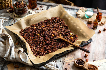 homemade baked chocolate granola with nuts, oat flakes on baking tray with parchment stands on grey wooden table
