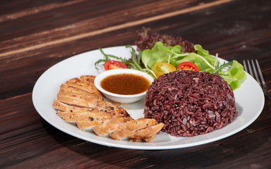 Top view of roasted chicken breast fillet steak with brown rice/coarse rice, vegetables salad and spicy sauce in white plate on wooden table. Healthy food menu, eating right concept.