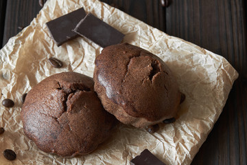 Two homemade chocolate muffins on parchment with coffee beans and pieces of chocolate. Dark wooden background, closeup, selective focus