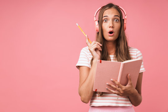 Image of surprised beautiful girl writing down notes at diary book and listening to music with headphones