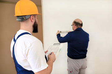 Engineer, contractor and project manager with his colleague, construction worker, handyman and builder is measuring wall room for renovation using spirit level tool, checking in apartment blueprint.