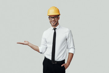 Handsome young man in helmet looking at camera and pointing copy space while standing against grey background