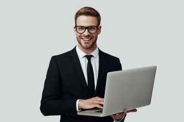 Handsome young man in formalwear working using laptop and smiling while standing against grey background