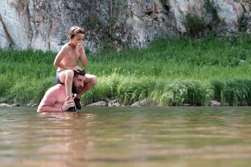 The son sits on the shoulders of a young father and holds his nose in his hands before jumping into the water. Swimming in a mountain river or lake. Camping lifestyle.
